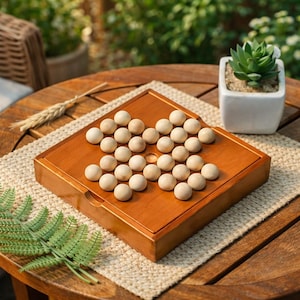 May include: A square wooden peg solitaire board game featuring a smooth, polished finish and a grid of circular indentations. The board is filled with light-colored, spherical wooden marbles, leaving only the center hole empty for gameplay.