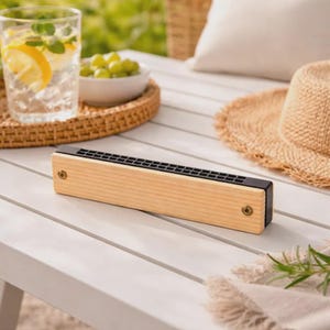 Peut inclure: Un harmonica rectangulaire doté d'un boîtier en bois naturel clair et d'une plaque supérieure en métal noir avec des trous d'aération visibles. L'instrument est fixé par deux petites vis couleur laiton sur la face avant, au design linéaire.