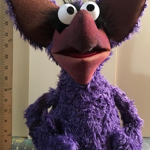 May include: A purple fuzzy puppet with large pointed ears, a dark red beak, and two white eyes with black pupils. A wooden ruler is positioned vertically on the left side, showing the puppet stands approximately 20 inches tall from base to ear.