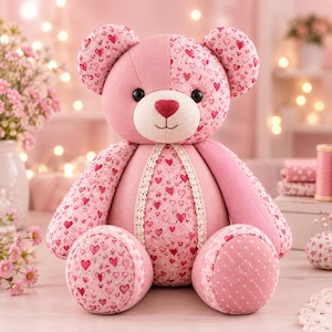 Pu&ograve; includere: Un orsacchiotto di peluche realizzato con vari tessuti rosa con motivi a cuore e sezioni rosa tinta unita. L'orso presenta occhi a bottone neri, un naso a forma di cuore rosa scuro e un bordo in pizzo color crema lungo il centro del busto.