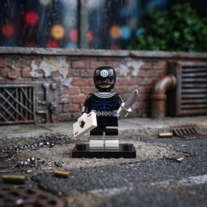 Peut inclure: Une figurine en plastique noir et blanc portant un masque avec un symbole de cible sur le front. Le personnage présente un motif bleu sur la poitrine, tient une carte à jouer blanche avec un pique dans une main et un couteau jouet argenté.