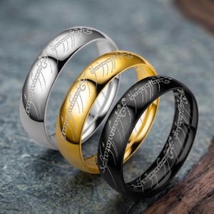 May include: Three metal rings in silver, gold, and black tones, each featuring a polished finish and an engraved script pattern that wraps around the entire outer band. The rings are arranged in a staggered, overlapping formation.