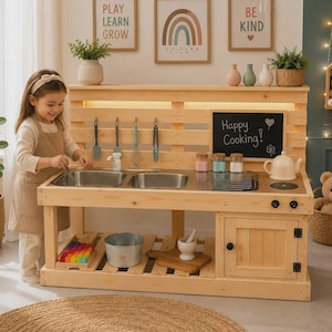 Puede incluir: Una cocina de juguete de madera con un fregadero doble de acero inoxidable, una pizarra negra con el texto Happy Cooking, un armario con un pomo negro, utensilios colgantes, tres frascos pequeños, una tetera blanca y un cubo de metal.
