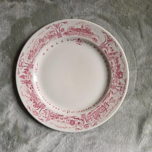 May include: A white ceramic plate with a red decorative border featuring illustrations and text. The plate has the words "HOWARD JOHNSON'S RESTAURANTS" around the edge. The plate is round and has a plain white center.