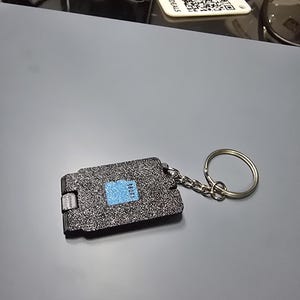 Könnte beinhalten: Ein rechteckiger schwarzer Schlüsselanhänger mit einer texturierten, glitzernden Oberfläche und einem kleinen blauen quadratischen Akzent in der Mitte. Der Schlüsselanhänger verfügt über einen silberfarbenen Metallring an einer kurzen Kette.