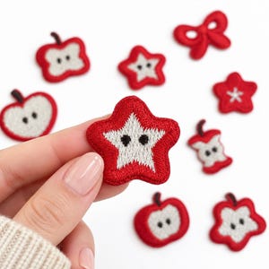 May include: A collection of embroidered fabric patches featuring red and white designs, including star shapes, apple slices, and a bow. The patches have a textured, stitched appearance with small black details, showcasing various fruit-themed motifs.