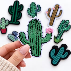May include: A collection of embroidered cactus patches in various shapes and shades of green, brown, and teal. One patch features a bright pink flower on a green cactus arm. The patches have white borders and are designed for fabric application.