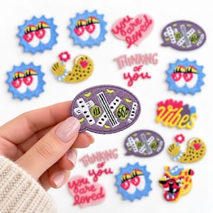 May include: A collection of embroidered patches featuring various designs including blue starbursts with eyes, pink hearts with the text you are loved, purple speech bubbles with white patterns, yellow leopard print shapes, and pink text reading thinking of you.