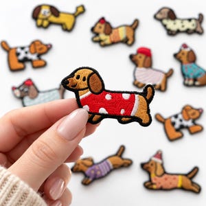 May include: A collection of embroidered dachshund patches featuring various dogs in colorful sweaters and hats. One patch in the foreground shows a brown dachshund wearing a red sweater with white polka dots, while others show dogs in different outfits.