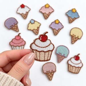 May include: A collection of embroidered fabric patches featuring various ice cream cones and cupcakes in pastel colors like pink, blue, yellow, and lavender, with some topped with small yellow stars or red cherries.