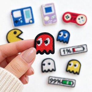May include: A collection of embroidered patches featuring video game themes, including a yellow circular character, a red ghost, a blue ghost, a white ghost, a red game controller, handheld consoles, and battery life indicators labeled 1% and 99%.