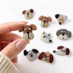 May include: A collection of small, embroidered dog face patches in various breeds and colors, including shades of brown, gray, white, and black. Each patch features detailed stitching to define the eyes, nose, and ears of the different canine designs.