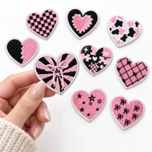May include: A collection of nine heart-shaped embroidered patches featuring various patterns including black and pink checkerboard, cow print, stars, bows, and puzzle pieces, with one patch held up showing a black and pink sunburst design.