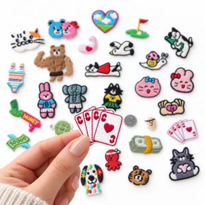 May include: A collection of small embroidered fabric patches featuring various colorful designs including animals, playing cards, money, and food items, with one hand holding a patch depicting three playing cards with red hearts.
