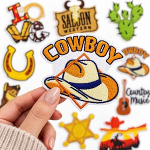 May include: An embroidered fabric patch featuring a cream-colored cowboy hat with a blue band, set against a white background with the orange text COWBOY arched above. The patch is shaped like a diamond with an orange border.