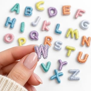May include: A collection of small, individual embroidered alphabet letter patches in various pastel colors including blue, yellow, pink, purple, and orange. Each letter features a raised, stitched texture with a defined border.
