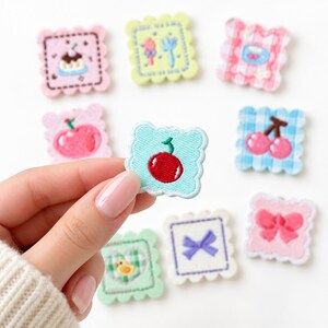 May include: A collection of square embroidered patches with scalloped edges, featuring various designs including a cherry, a bow, a duck, a pudding, and a teacup in pastel colors like pink, blue, green, and yellow on a white background.