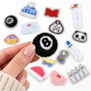 May include: A collection of small embroidered patches featuring various designs including an eight ball, a blue winter hat, a red book, a snowman, a panda face, a game controller, a yellow skateboard, a pink hat, and pink underwear.