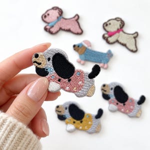 May include: A collection of embroidered dog patches featuring various breeds and colors. One patch shows a dog with a pink polka-dot sweater, black ears, and a tan face. Other patches include a blue sweater-wearing dog, a pink dog, and a yellow sweater dog.