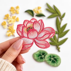 May include: An embroidered lotus flower patch in shades of pink and white with a green and yellow center. Additional embroidered patches are visible in the background, including yellow floral sprigs, a green bamboo stalk, and two small green lily pads.