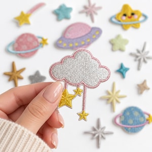 May include: A collection of embroidered space-themed patches including a silver cloud with hanging yellow stars, planets with rings, various stars, and a purple UFO. The patches feature vibrant colors like pink, blue, yellow, and silver.