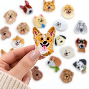 May include: A collection of small, embroidered dog face patches in various breeds and colors. One patch featuring a tan and white Corgi with a pink tongue is held in the foreground, while other assorted dog head patches are scattered around.