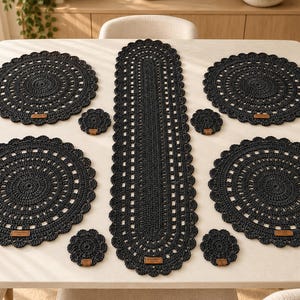 May include: A set of black crocheted table linens including four round placemats, four small round coasters, and one long oval table runner. Each piece features a scalloped edge and a small rectangular brown leather-like brand tag.