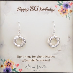 May include: Silver earrings with a circle design made of eight intertwined rings. The earrings are displayed on a card with the text "Happy 80th Birthday" and "Eight rings for eight decades of beautiful memories!"  The card also includes the name "Alma Valle jewelry".
