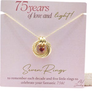 May include: A gold-colored necklace with a pendant featuring seven interlocking rings, one with a small red gemstone. The necklace is displayed on a pink card with the text "75 years of love and light!" and "Seven Rings".