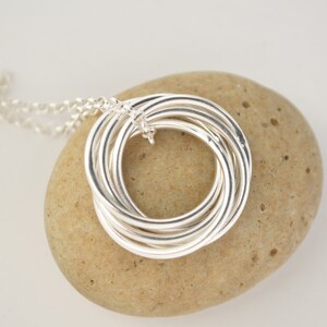 May include: Silver necklace with a pendant made of multiple intertwined rings. The pendant is resting on a light brown stone.