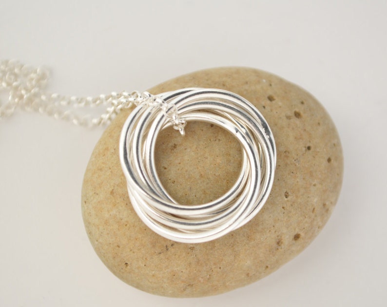 May include: Silver necklace with a pendant made of multiple intertwined rings. The pendant is resting on a light brown stone.