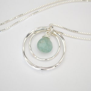 May include: A silver necklace with a delicate chain and a pendant featuring three concentric circles with a light blue gemstone in the center.