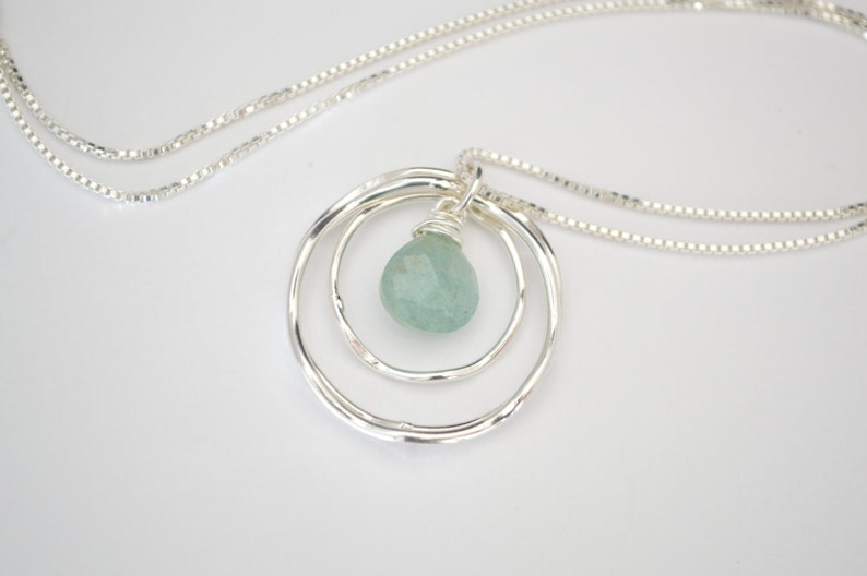 May include: A silver necklace with a delicate chain and a pendant featuring three concentric circles with a light blue gemstone in the center.