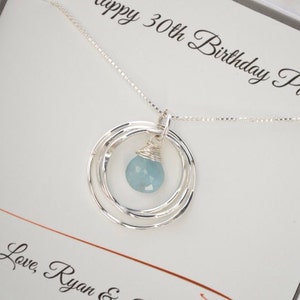May include: A silver necklace with a blue gemstone pendant and three silver rings. The necklace is in a white box with the text "Happy 30th Birthday Pam! Love, Ryan &"