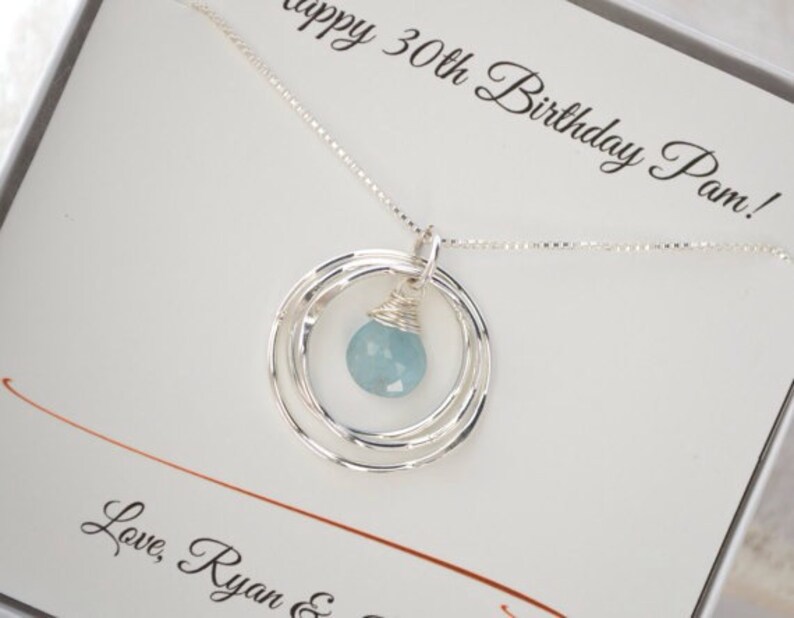 May include: A silver necklace with a blue gemstone pendant and three silver rings. The necklace is in a white box with the text "Happy 30th Birthday Pam! Love, Ryan &"