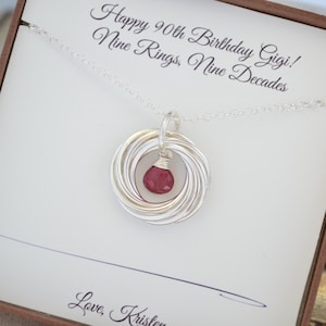 以下が含まれることがあります： 9つのインターロッキングリングと赤い宝石のペンダントが付いたシルバーネックレス。ギフトボックスには「Happy 90th Birthday Gigi! Nine Rings, Nine Decades.」と書かれています。