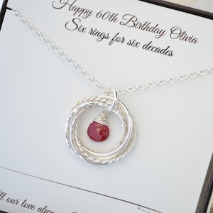 May include: A silver necklace with six interlocking rings and a red gemstone pendant. The necklace is presented in a gift box with the text "Happy 60th Birthday Olivia Six rings for six decades."