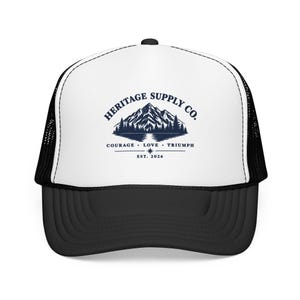 May include: A white and black trucker hat featuring a dark blue graphic of a mountain range above a lake. The text reads HERITAGE SUPPLY CO. with the words COURAGE, LOVE, TRIUMPH and EST. 2026 printed below the mountain illustration on the front panel.