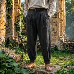 May include: High-waisted charcoal gray trousers featuring a wide waistband with a vertical row of five buttons, deep front pleats, and tapered legs with cuffed hems, paired with a long-sleeved beige shirt and brown leather shoes.
