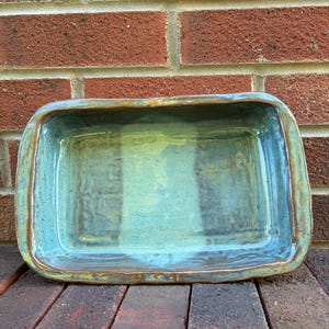 May include: A rectangular ceramic baking dish with a glossy glaze in shades of teal, blue, and green. The vessel features an uneven, hand-formed rim with visible brown clay edges and a smooth, reflective interior surface with subtle color variations.