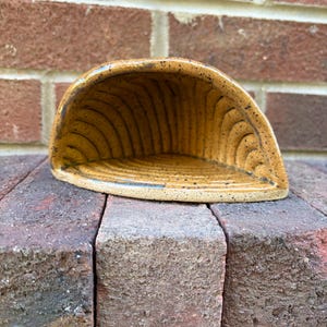 May include: A semi-circular ceramic wall pocket with a speckled mustard yellow glaze. The interior features a series of carved, radiating grooves that extend from the base to the curved outer edge, creating a textured, shell-like appearance.