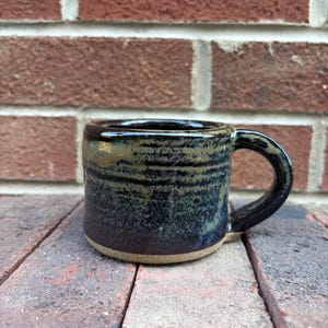 May include: A ceramic mug with a cylindrical shape and a curved handle. The exterior features a dark, mottled glaze with deep blue and black tones, transitioning to a lighter, earthy tan color at the unglazed base of the vessel.