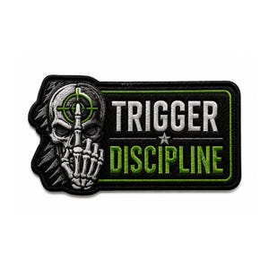 May include: A rectangular embroidered patch with a black background and a green border. On the left, a grey skeletal hand makes a gesture in front of a skull with a green crosshair. The text TRIGGER DISCIPLINE is embroidered in white and green.