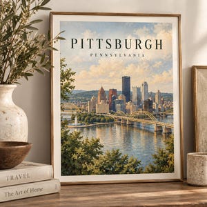 Op de afbeelding: Een ingelijste kunstdruk met een geschilderd stadsgezicht van Pittsburgh, Pennsylvania. De afbeelding toont de skyline, een rivier, een gele brug en een fontein onder een bewolkte lucht. De tekst PITTSBURGH PENNSYLVANIA staat bovenaan.
