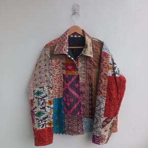 May include: A long-sleeve jacket constructed from various patchwork fabric panels featuring diverse patterns and colors including red, blue, purple, and cream. The garment has a collared neckline, a button-down front, and visible quilted stitching.