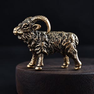 Puede incluir: Una pequeña figura de latón de un carnero de perfil. La escultura de metal presenta un pelaje texturizado, cuernos curvados y un acabado de estilo antiguo con reflejos oscuros en las hendiduras de la superficie metálica.