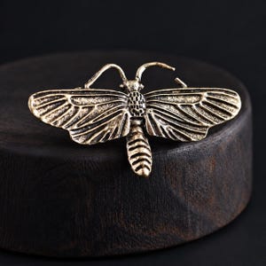 May include: A metallic moth brooch with an antique brass finish, featuring detailed wing patterns, a textured thorax, and a segmented abdomen. The insect design includes two curved antennae extending from the head, showcasing a weathered appearance.