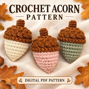 Op de afbeelding: Drie handgehaakte eikels met gestructureerde bruine dopjes en gladde lichamen in crème, lichtroze en saliegroen. De tekst CROCHET ACORN PATTERN en DIGITAL PDF PATTERN is zichtbaar. De eikels liggen op een lichte stof met herfstbladeren.