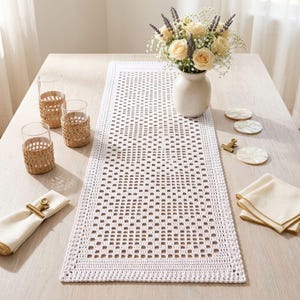 May include: A rectangular white crochet table runner with a geometric grid pattern. The runner is accompanied by two glass tumblers with woven seagrass sleeves, a ceramic vase with cream roses and lavender, three round coasters, and napkins.