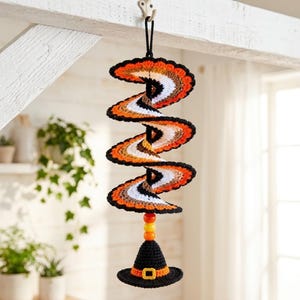 May include: A hanging crochet spiral decoration featuring alternating stripes of black, white, orange, and brown yarn. The spiral is suspended by a black loop and terminates with three round orange beads above a small black crochet witch hat.